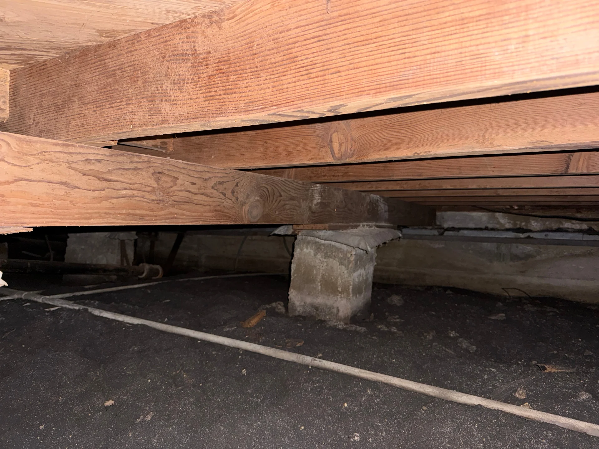 Wooden beams and concrete pier supports visible in a crawlspace beneath a pier-and-beam home